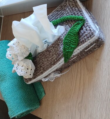 Gehäkelte Taschentuchbox-Hülle in Braun mit weißen Blüten und grünen Blättern, neben einem türkisfarbenen Handtuch
