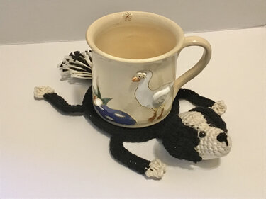 Beigefarbene Tasse mit Gänsemotiv auf gehäkeltem Untersetzer in Hundeform.