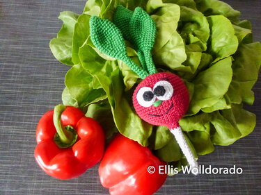 Gehäkelter roter Rettich mit Gesicht auf grünen Salatblättern neben zwei roten Paprika
