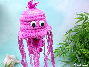 Pinkfarbene gehäkelte Qualle mit großen Wackelaugen und langen Garntentakeln neben grünen Pflanzenblättern