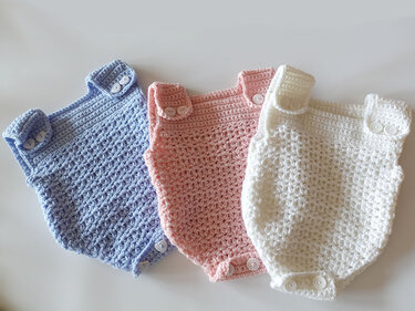 Drei gehäkelte Baby-Strampler in Hellblau, Rosa und Weiß nebeneinander mit Knöpfen an Schultern und Schritt.