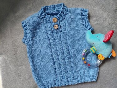 Hellblaue gestrickte ärmellose Babyweste mit mittigem Zopfmuster und zwei Holzknöpfen, liegend neben einer Elefanten-Rassel