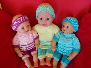 Drei Babypuppen in gestrickten Outfits mit Stirnbändern in Rosa, Gelb-Grün und Türkis auf rotem Sessel