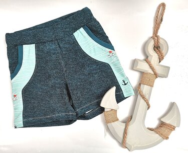 Dunkelblaue Kinder-Shorts mit hellblauen, maritim gemusterten Seitenpartien, daneben ein weißer Deko-Anker.