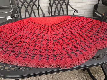 Rotes gehäkeltes Halbrundtuch mit feinem Lochmuster, ausgebreitet auf einem dunklen Gartentisch.