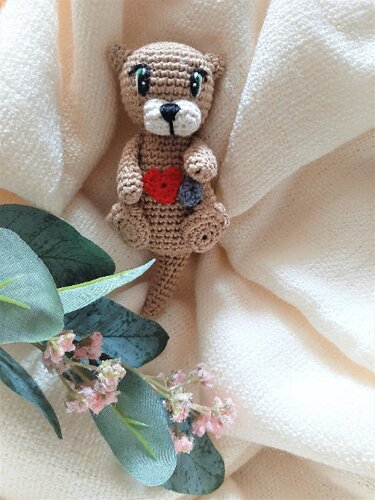 Beigefarbener gehäkelter Fischotter mit rotem Herz und grauer Blume auf hellem Stoff