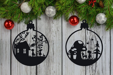 Zwei schwarze Silhouetten in Form von Weihnachtskugeln: links Haus mit Kindern und Bäumen, rechts Pilze mit Reh und Kind, vor Holzuntergrund und Tannenzweigen.