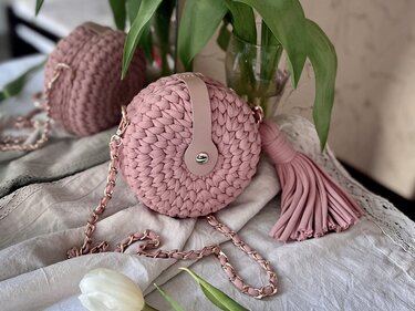 Pink round crocheted shoulder bag with tassel, woven chain strap, and flap with metal snap, placed on light fabric beside a white tulip.
