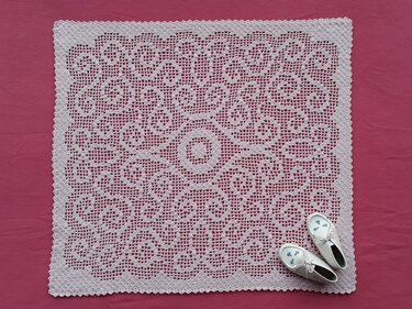 Quadratische weiße Filet-Häkeldecke mit zentralem Rundmotiv auf rosa Hintergrund, daneben weiße Babyschuhe.