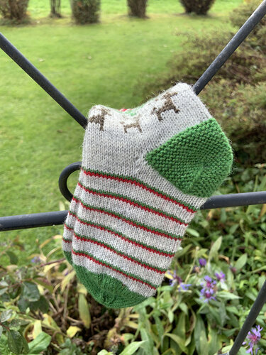 Gestrickte hellgraue Kindersocke mit roten Streifen, Rentiermotiven und grüner Ferse, aufgehängt an Metallgeländer