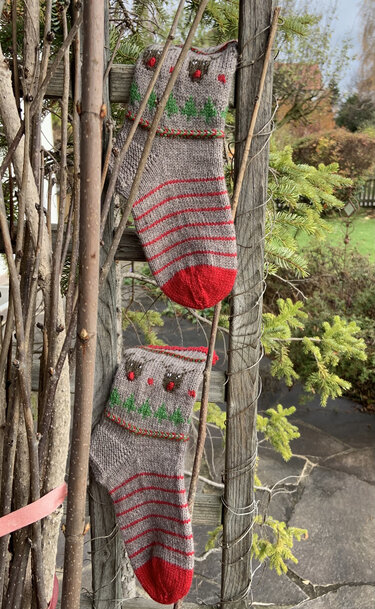 Zwei gestrickte graue Weihnachtssocken mit roten Streifen, roten Spitzen und Rentier‑/Tannenbaum‑Motiv an einem Holzspalier.