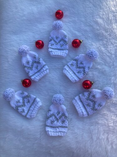 Sieben weiße gestrickte Mini-Mützen mit Bommel und silbernem Zickzackmuster auf weißem Plüsch, dekoriert mit roten Kugeln
