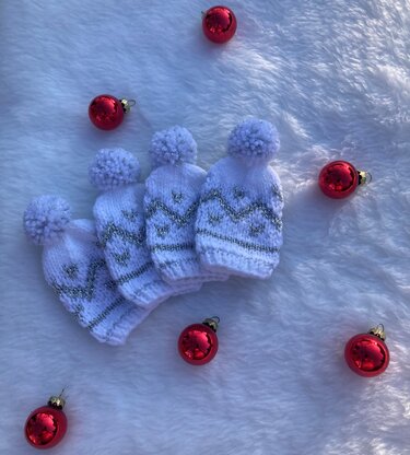 Vier weiße, gestrickte Mini-Mützen mit silbernem Muster und Pompons auf weißem Kunstfell, umgeben von roten Weihnachtskugeln.