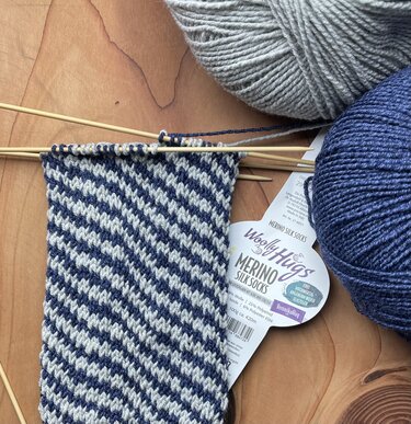Halbfertige gestrickte Socke mit diagonalen dunkelblauen und hellgrauen Streifen auf Nadelspiel, zwei Garnknäuel und eine Etikette auf Holzuntergrund