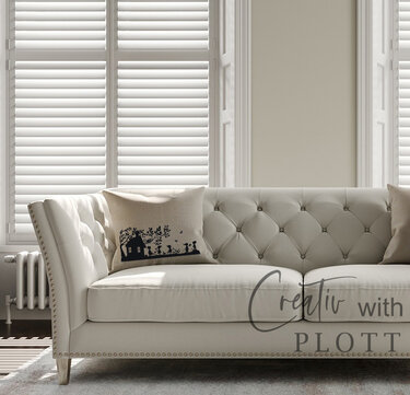 Beiges Zierkissen mit schwarzer Haus-Silhouette, Bäumen sowie Kinder- und Tierfiguren auf einem hellen Sofa.