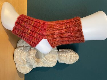 Weißer Schaufensterfuß mit rostroter, kurz gestrickter Beinstulpe mit Rippenmuster vor dunklem Hintergrund.