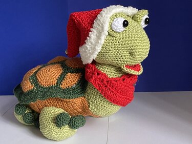 Gehäkelte Amigurumi-Schildkröte mit rot-weißer Weihnachtsmütze und rotem Schal vor blauem Hintergrund