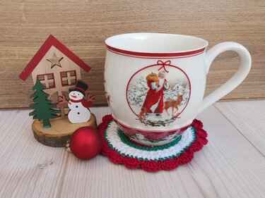 Gehäkelter runder Untersetzer in Rot, Weiß und Grün unter einer Tasse mit Weihnachtsmotiv, daneben Holzhaus, Schneemannfigur und rote Kugel