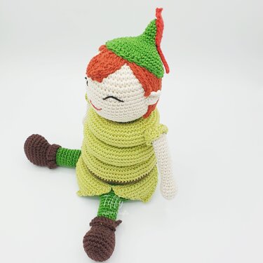 Amigurumi boy doll with orange yarn hair, green tunic, pointed hat with red feather, and brown boots in a sitting pose