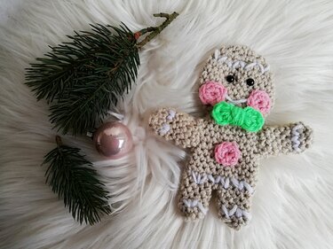 Kleiner gehäkelter Lebkuchenmann mit weißen Ziernähten, rosa Akzenten und grüner Fliege neben Tannenzweig und rosafarbener Kugel auf weißem Fell