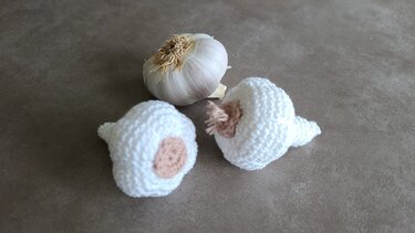 Zwei weiße, gehäkelte Knoblauchknollen mit beigefarbenen Zentren neben einer echten Knoblauchknolle