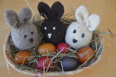 Heukorb mit drei gestrickten Hasen-Eierwärmern (grau, dunkelbraun, weiß) und bunten Ostereiern
