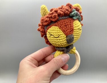 Hand hält gehäkelten Baby‑Greifling mit Tiergesicht und Holzring.