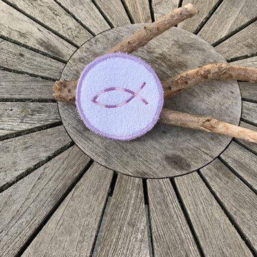 Runder, lilafarbener Untersetzer mit gesticktem Fischsymbol auf hölzerner Oberfläche