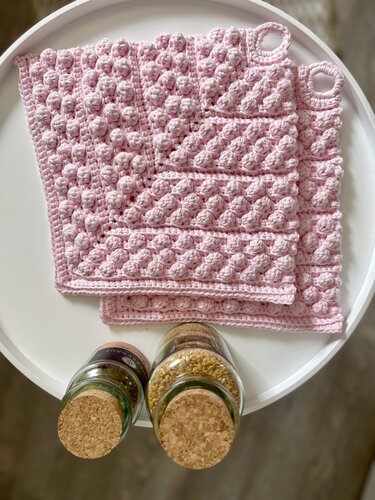 Zwei rosa gehäkelte Topflappen mit Noppenmuster und Aufhängeschlaufen auf einer weißen Ablage, daneben Gewürzgläser mit Korkdeckeln.