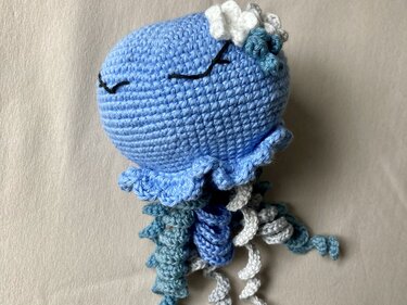 Gehäkelte Amigurumi-Qualle in Hellblau mit Rüschenrand, bestickten Augen und mehrfarbigen Spiral-Tentakeln.