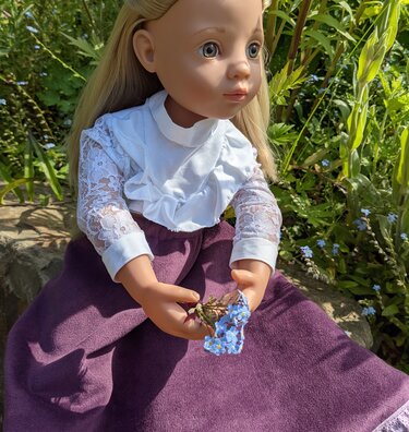 Nahaufnahme einer blonden Puppe mit weißer Bluse, transparenten Spitzenärmeln und lila Rock, sie hält kleine blaue Blumen