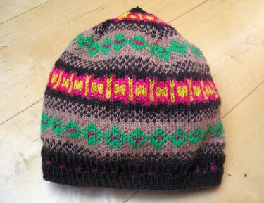 Gestrickte Beanie in Braun mit grünen, pinken und gelben Mustern auf hellem Holzuntergrund