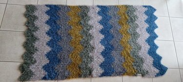 Gehäkelter Hochflor-Teppich mit Zickzackmuster in Blau, Grau und Senfgelb auf hellem Fliesenboden.