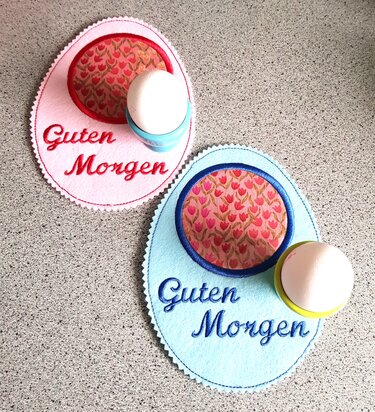 Zwei eiförmige, bestickte Filz-Platzsets in Rosa und Hellblau mit dem Text ‚Guten Morgen‘, rundem Tulpenstoff als Applikation und jeweils einem Eierbecher mit Ei.
