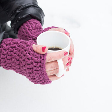 Hände mit lila gehäkelten, fingerlosen Armstulpen halten eine weiße Tasse mit dunklem Getränk vor verschneitem Hintergrund.