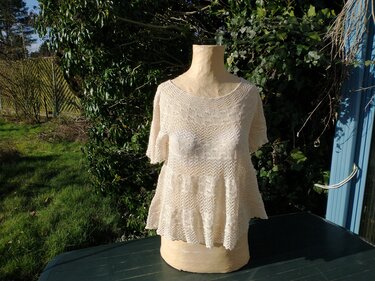 Cremefarbenes, gestricktes Damen‑Shirt mit kurzen Ärmeln und ausgestelltem Saum auf einer Schneiderpuppe vor Pflanzen.