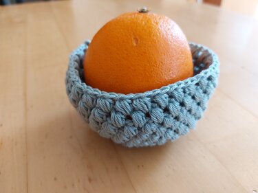 Orange in einem kleinen helltürkisen gehäkelten Korb mit Puffmaschen auf einem Holztisch.