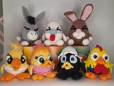 Sieben gehäkelte Amigurumi-Tiere: grauer Esel, weiße Fantasiefigur, brauner Hase, zwei gelbe Küken, dunkles Küken mit Eierschale und gelber Hahn.