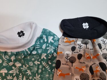 Zwei Kinderhosen mit Bündchen; links grün mit Pilz- und Blattmuster, rechts beige mit orangefarbenen Fuchs- und Baumillustrationen