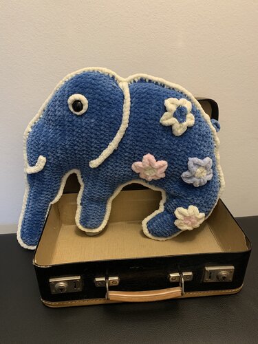 Blauer gehäkelter Amigurumi-Elefant mit weißer Umrandung und pastellfarbenen Blumen in einem offenen schwarzen Koffer