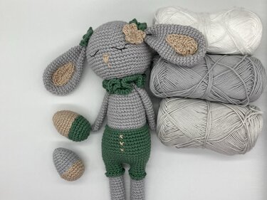 Gehäkelter grauer Amigurumi-Hase mit grüner Hose und zwei gehäkelten Eiern neben Garnknäueln
