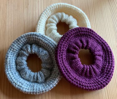 Drei gehäkelte Haargummis in cremeweiß, hellgrau und violett auf Holz