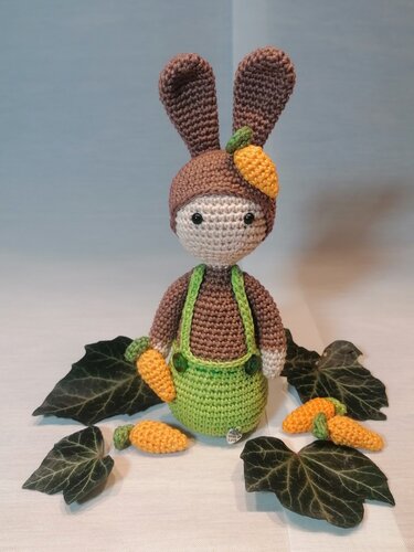 Gehäkelte Amigurumi-Hasenpuppe mit brauner Mütze, grüner Latzhose und kleinen gehäkelten Möhren.