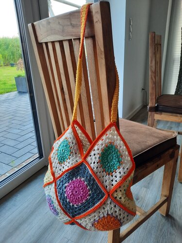 Gehäkelte Henkeltasche aus bunten Granny-Squares mit orangefarbener Umrandung an einem Holzstuhl.