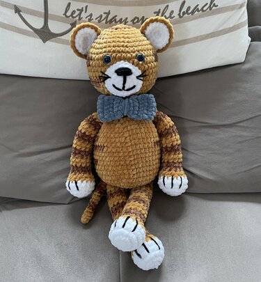 Gehäkelter Amigurumi-Tiger mit blau-grauer Fliege auf einem grauen Sofa.