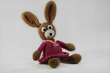 Sitzender gehäkelter Amigurumi-Hase in Braun mit langen Ohren und pinkem Häkelkleid vor hellem Hintergrund.