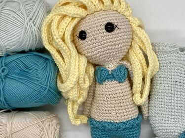 Gehäkelte Amigurumi-Puppe mit blondem Haar und türkisfarbenem Bikini, umgeben von Garnknäueln