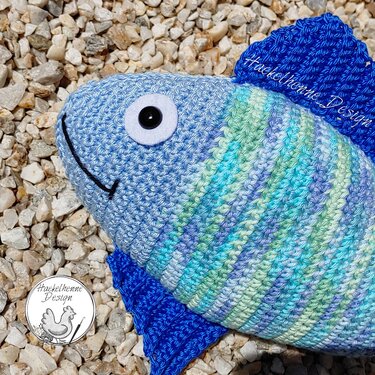 Nahaufnahme eines gehäkelten Fisches mit gestreiftem Körper, Filzauge und dunkelblauen Flossen auf Kies