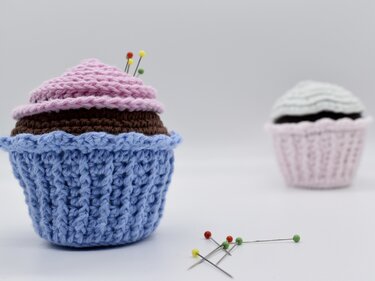 Gehäkeltes Cupcake-Nadelkissen mit Stecknadeln, weiteres Nadelkissen unscharf im Hintergrund