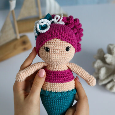 Gehäkelte Amigurumi-Meerjungfrauen-Puppe in Magenta und Türkis, von Händen gehalten; unscharfer Segelboot- und Korallenhintergrund.